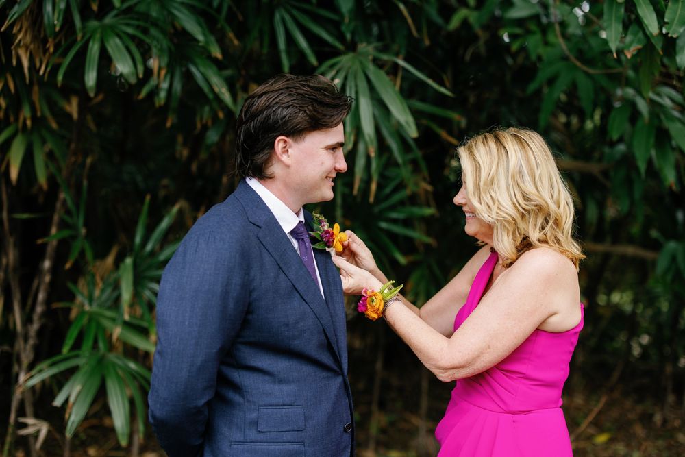 Woman in pink dress pinning boutonniere on man in navy suit outdoors with tropical greenery backdrop.