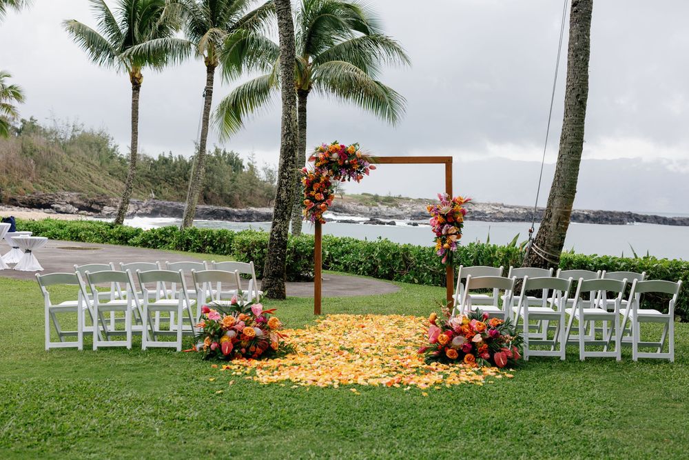 Tropical outdoor wedding ceremony setup with wooden arch, orange floral arrangements, white chairs, and ocean views in Hawaii.