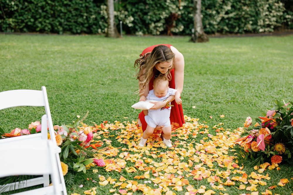 Woman in red dress bending down to help a young child walk along a flower petal aisle at an outdoor wedding.