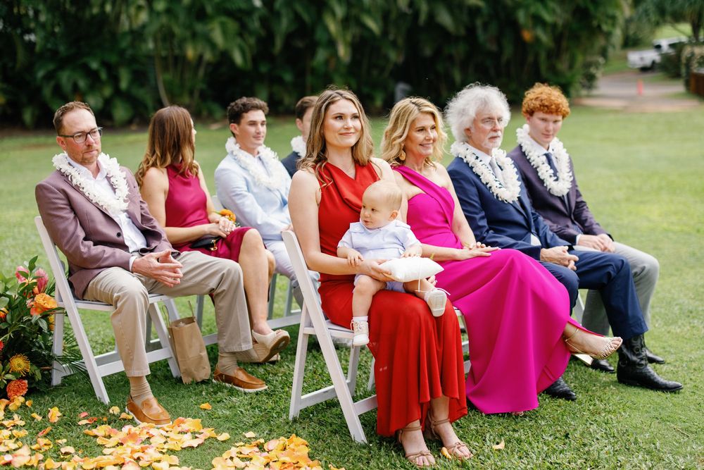 Family members in colorful formal attire seated on white chairs at an outdoor wedding ceremony on green lawn.
