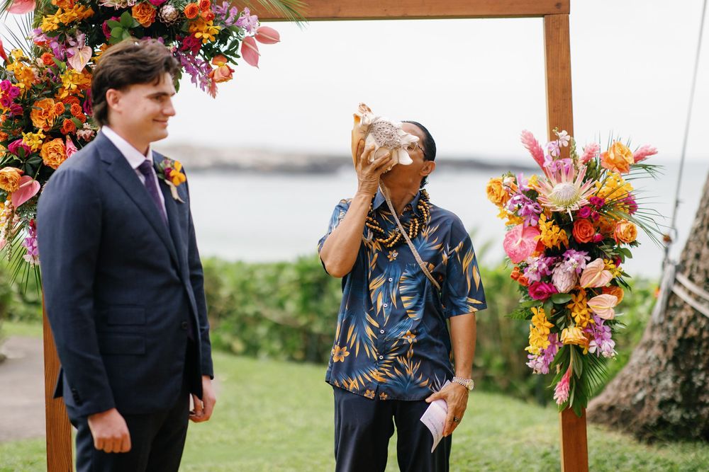 Officiant blows conch shell during outdoor Hawaiian wedding ceremony beside groom at floral arch.