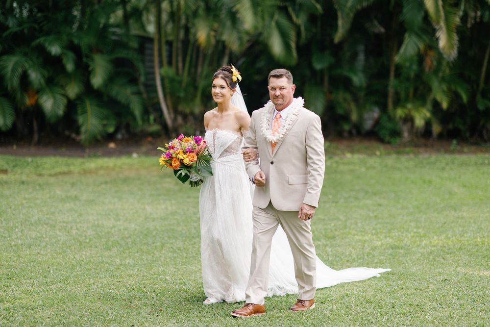 Bride and groom pose together on green lawn with tropical trees, bride holding vibrant bouquet in Hawaii wedding.