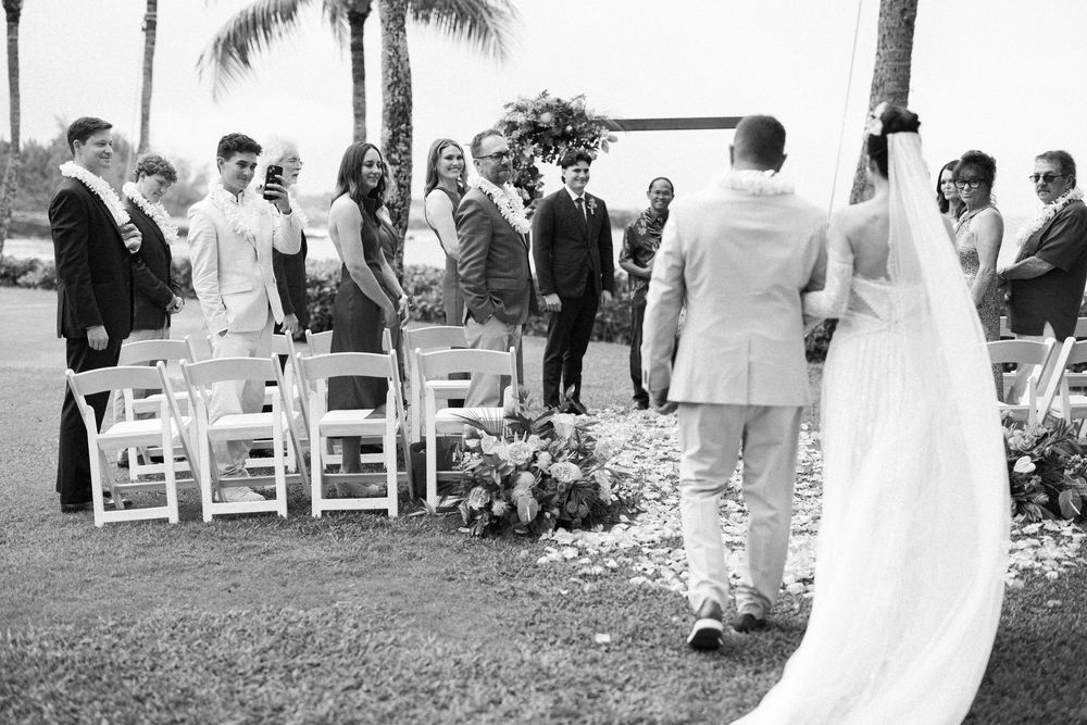 Bride and groom walk down the aisle at an outdoor wedding ceremony surrounded by guests on a sunny day.