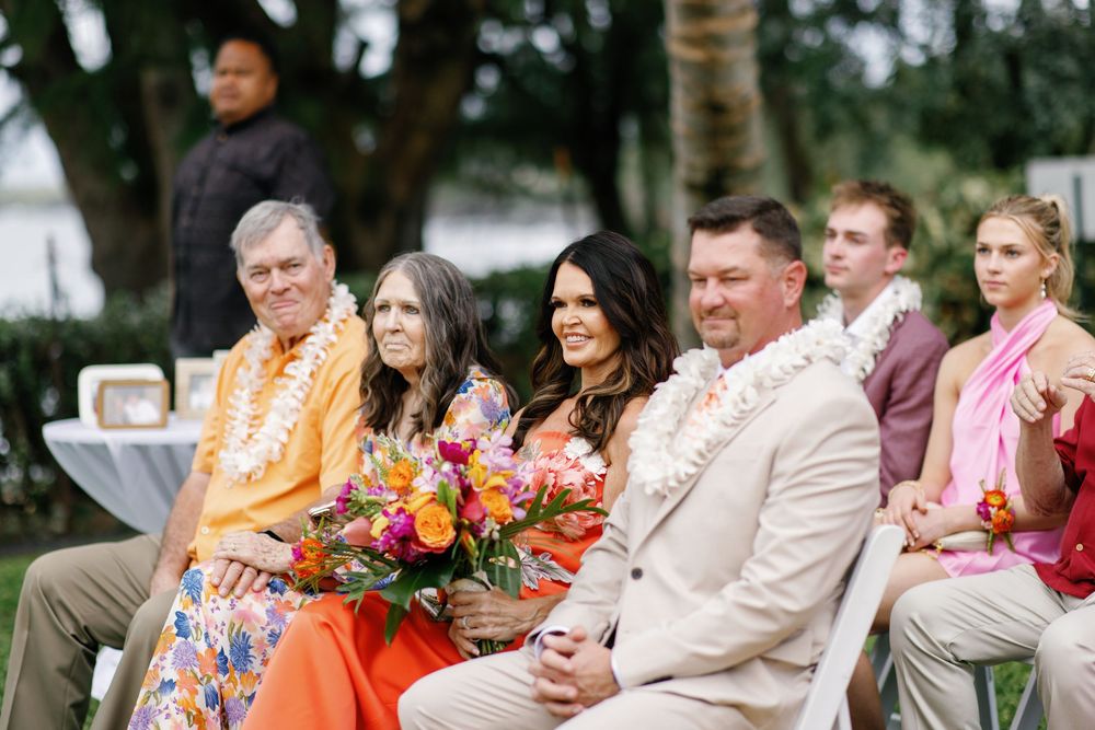 Wedding guests in colorful attire sit outdoors holding tropical bouquets during a Hawaii ceremony.