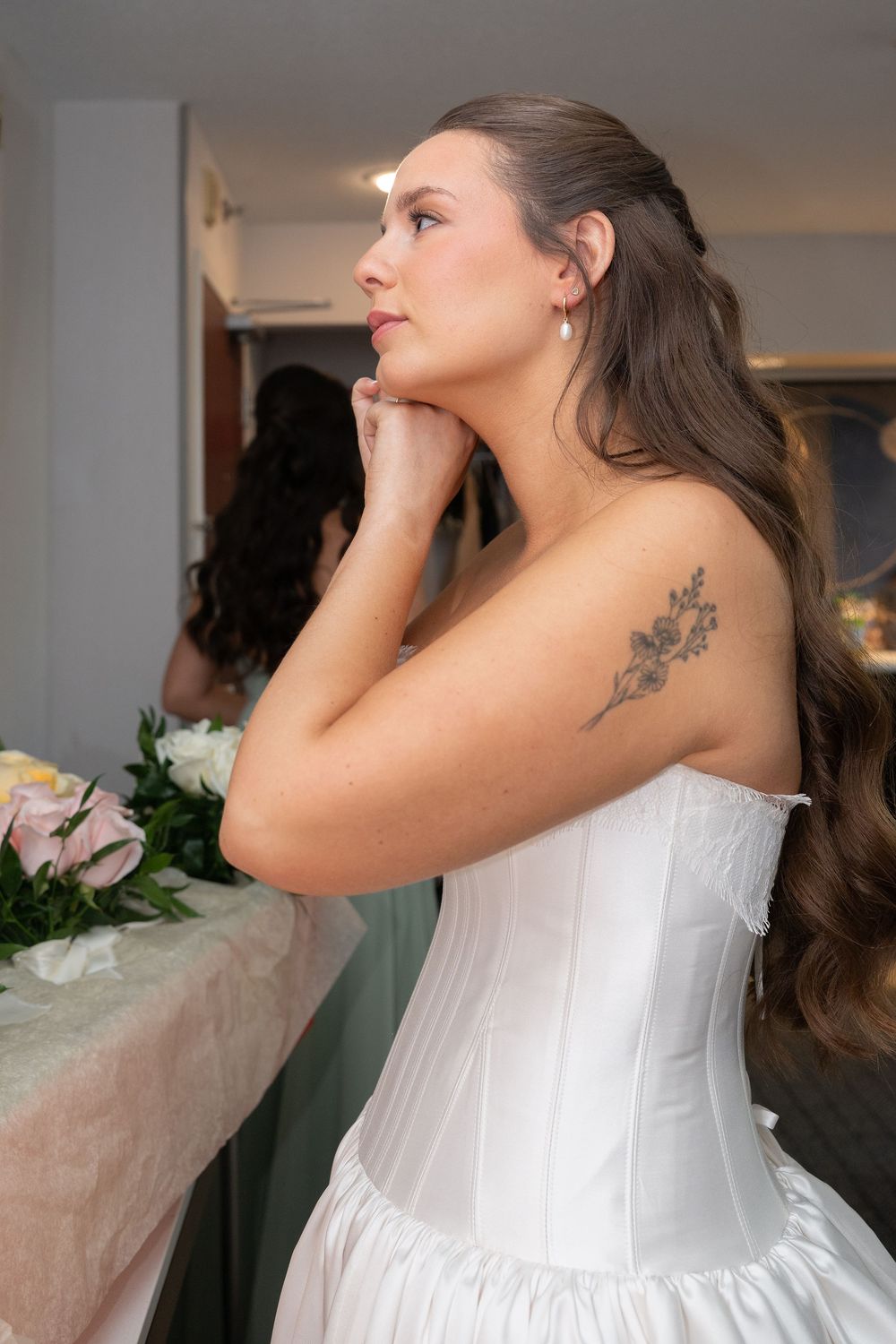 Bride in white strapless corset gown with floral tattoo, posing thoughtfully beside flowers in a bridal suite.