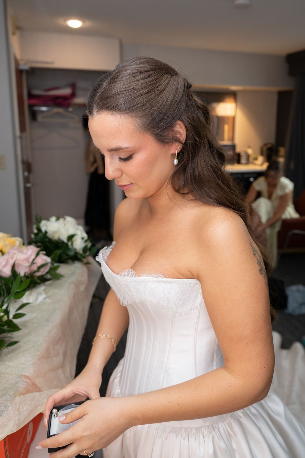 Bride in white strapless wedding gown holding phone, with flowers in background.