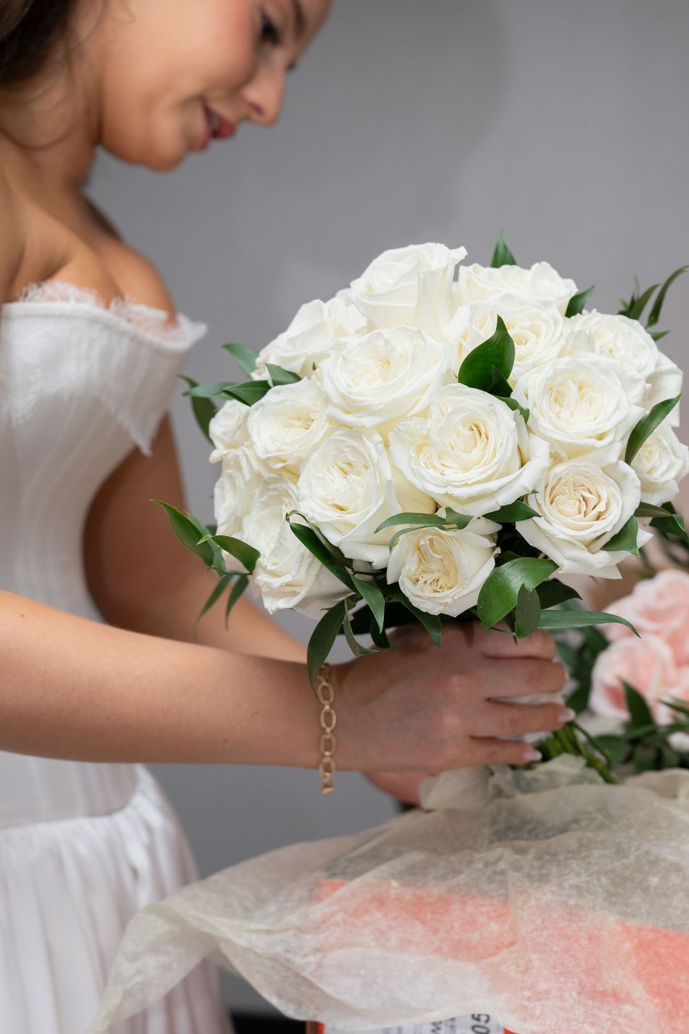 Bride in white dress holding elegant bridal bouquet of white roses with green leaves on gray background.