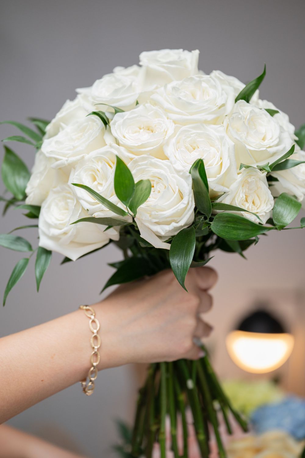 Hand holding a bridal bouquet of white roses with green foliage, gold bracelet visible, gray background.