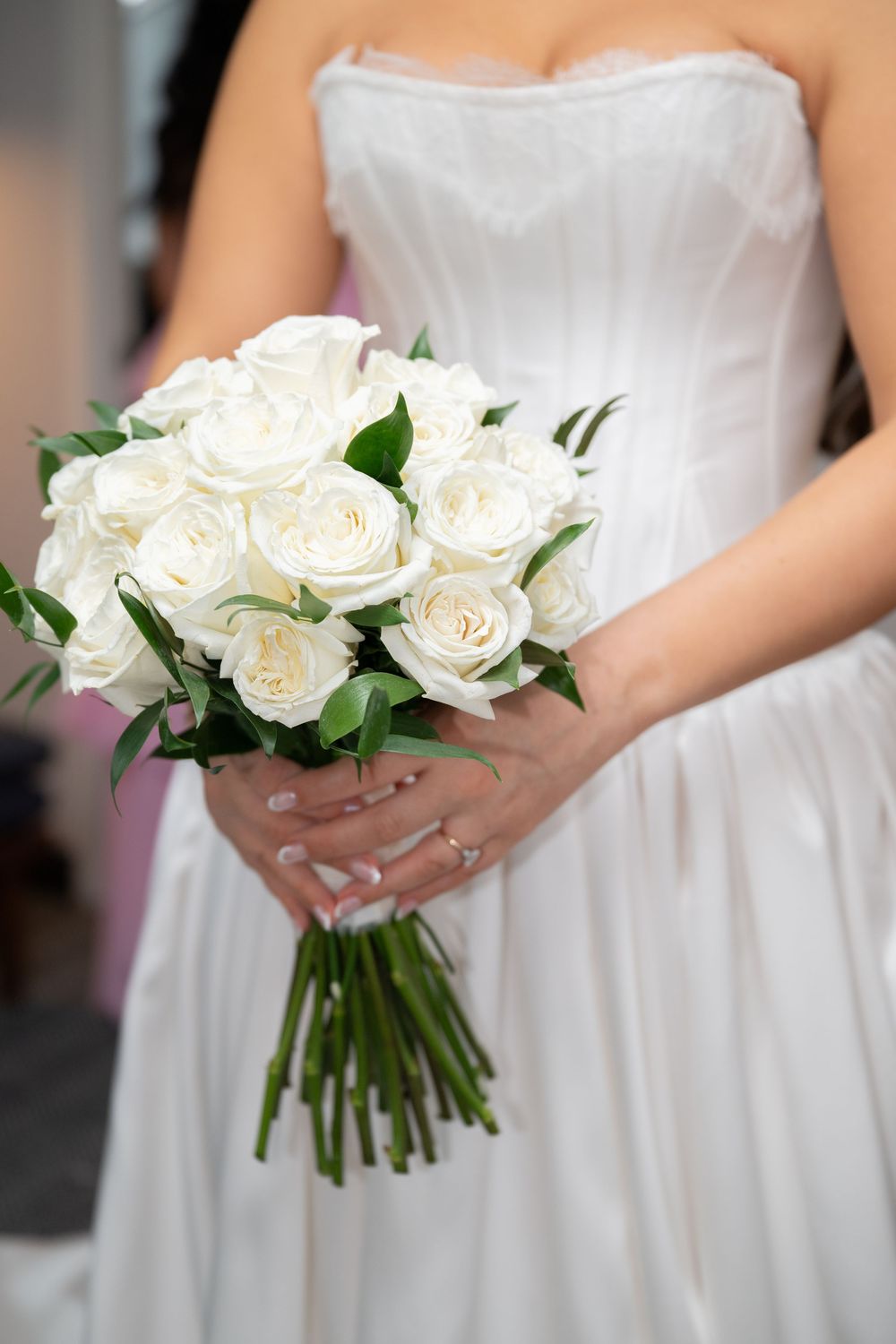 Bride in white strapless gown holding elegant bouquet of white roses with green stems at wedding ceremony.