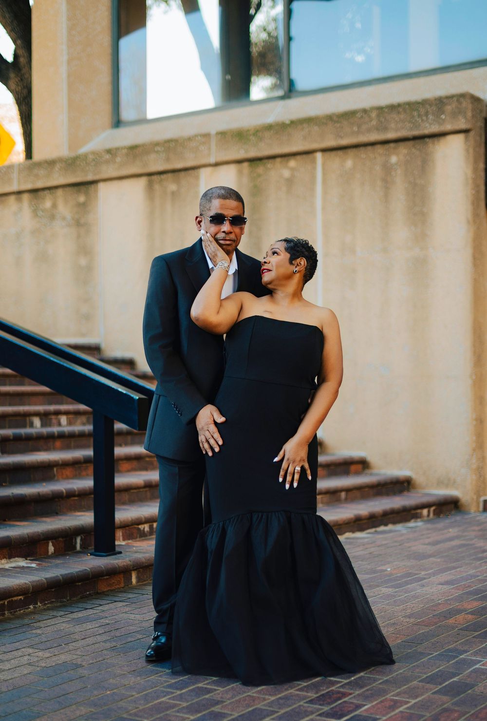 An elegant couple poses outdoors in matching black formal attire, with the woman in a strapless mermaid gown.
