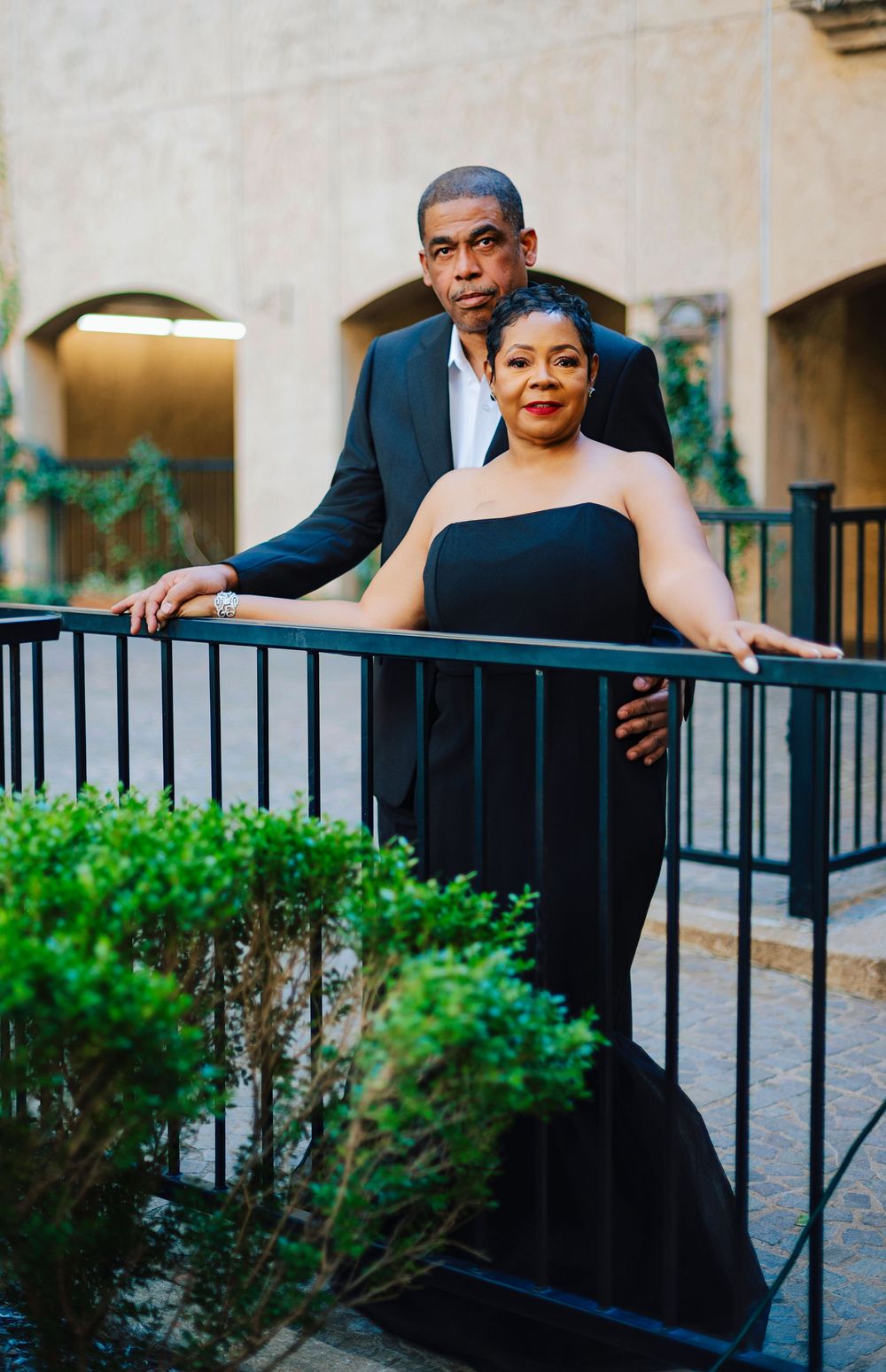 Elegant couple posing on a balcony railing, man in dark suit, woman in black strapless gown, surrounded by greenery.