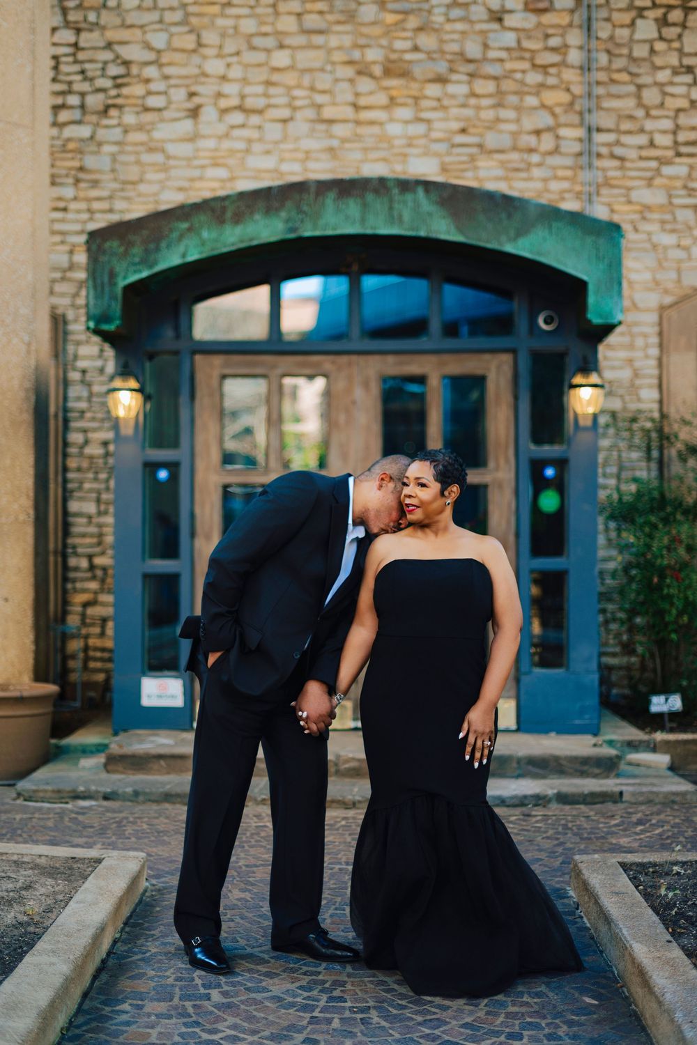 Man in black suit kissing woman in black mermaid gown outside stone building entrance.