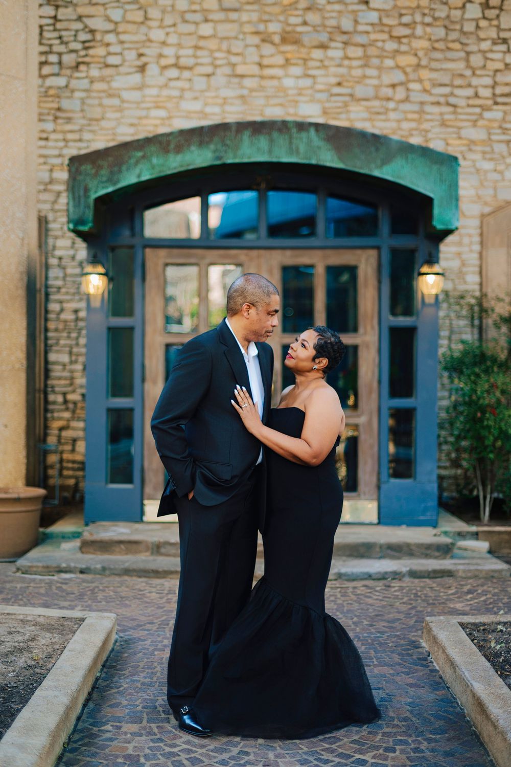 Couple in matching black formal attire sharing an intimate moment before a stone building entrance.