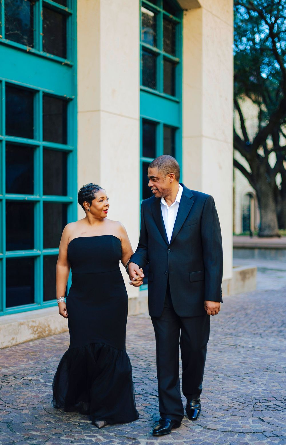 Stylish Black couple in formal black attire walking hand in hand past a teal-windowed building.