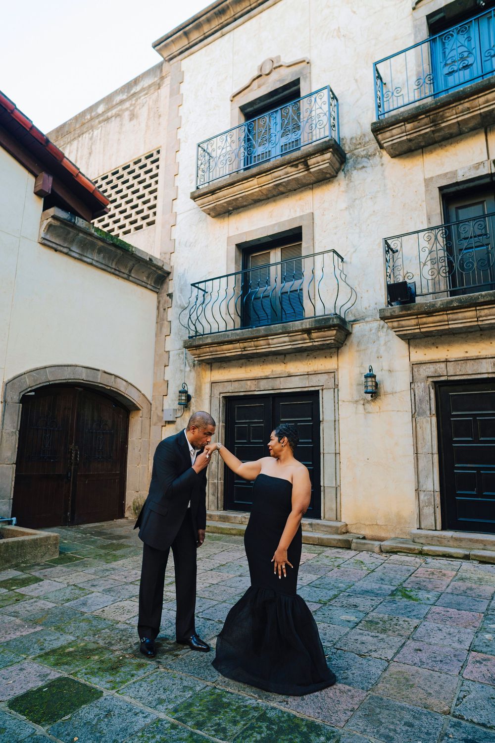 Man in black suit kisses womans hand in a historic cobblestone courtyard with wrought iron balconies.