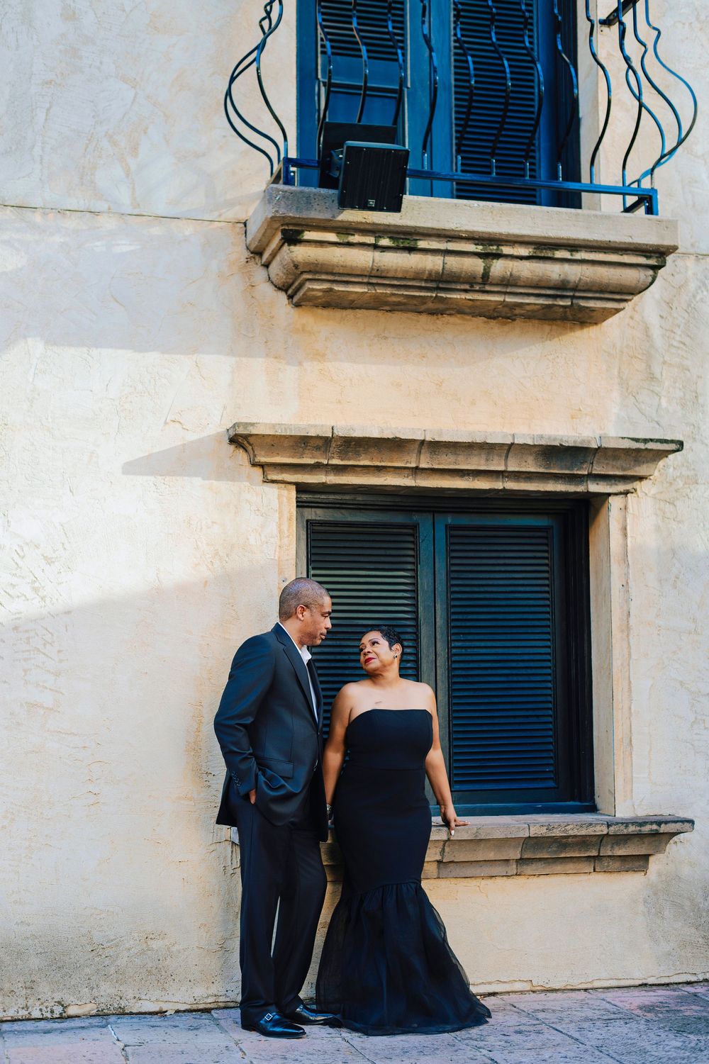 Elegant couple in matching black formal wear posing together against a historic stone building with blue shuttered windows.