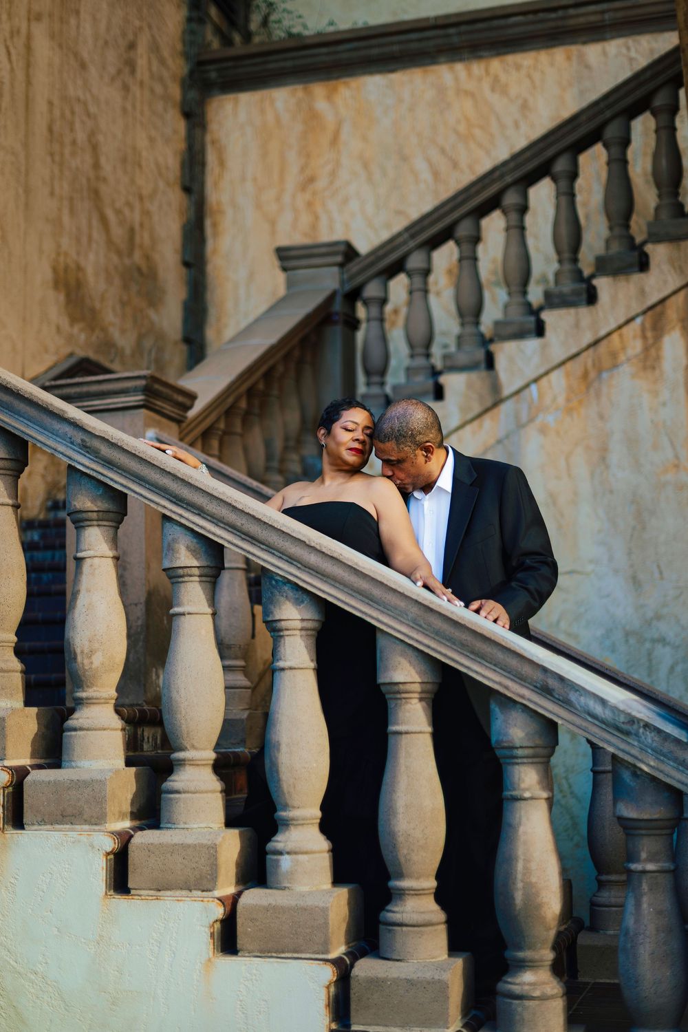 An elegant couple shares an intimate moment on ornate stone balustrade stairs at a classic architectural venue.