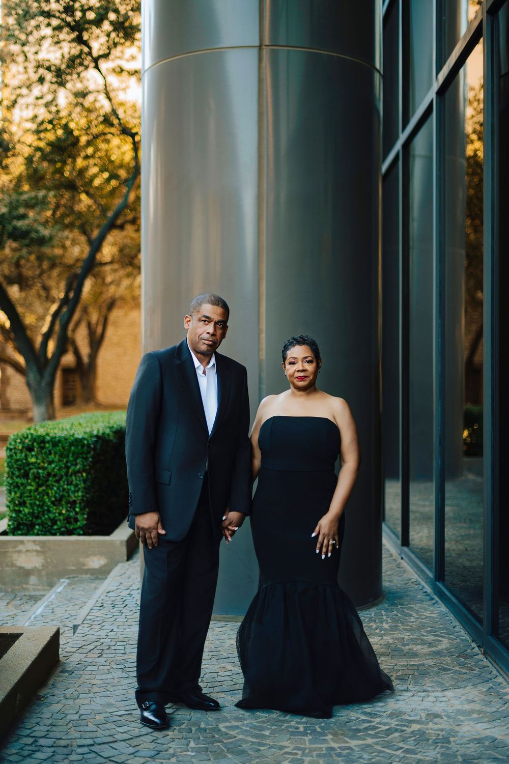 Elegant couple in matching black formal attire posing together outdoors near modern architectural columns.