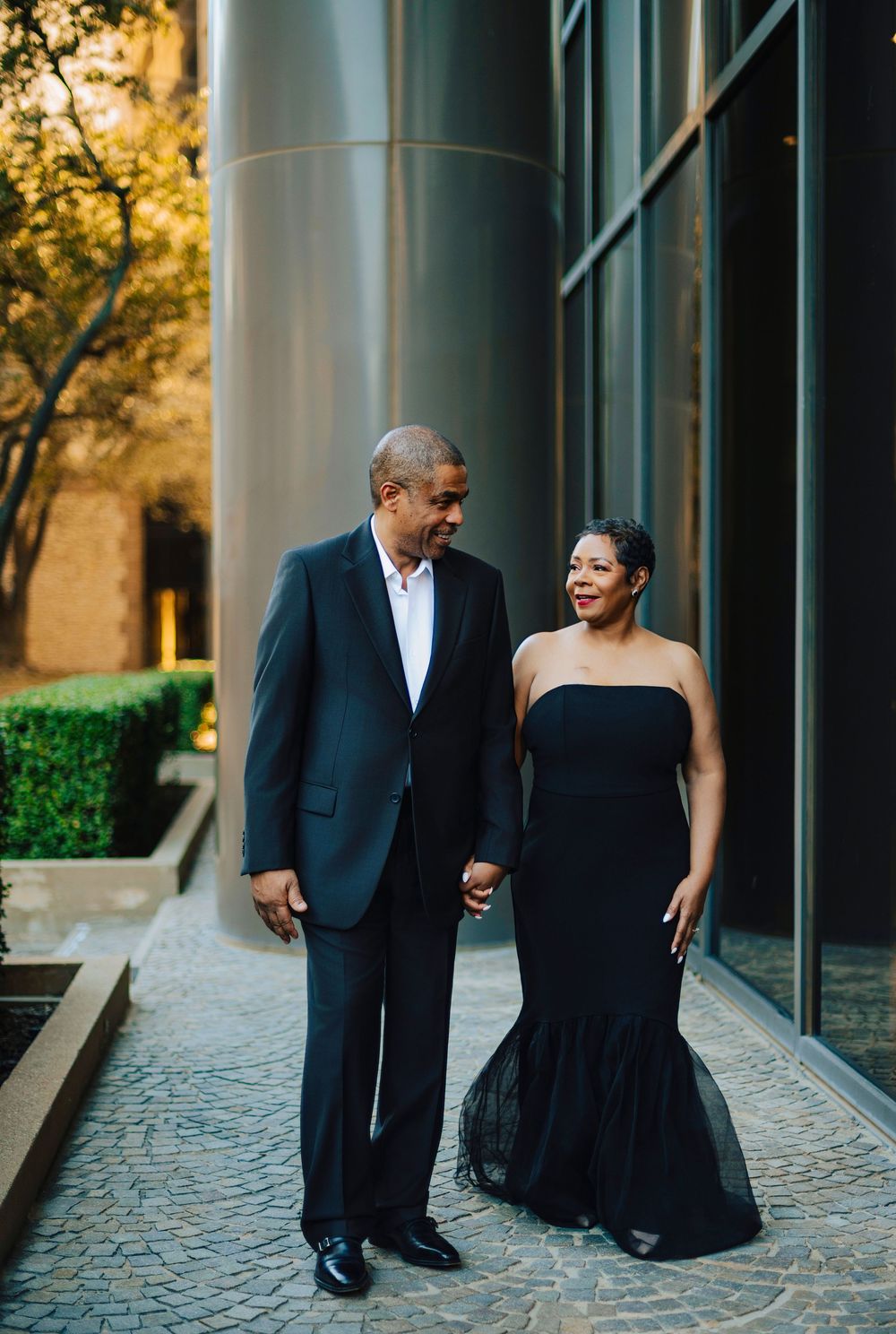 Stylish couple in black formal wear walking hand in hand along modern building walkway.