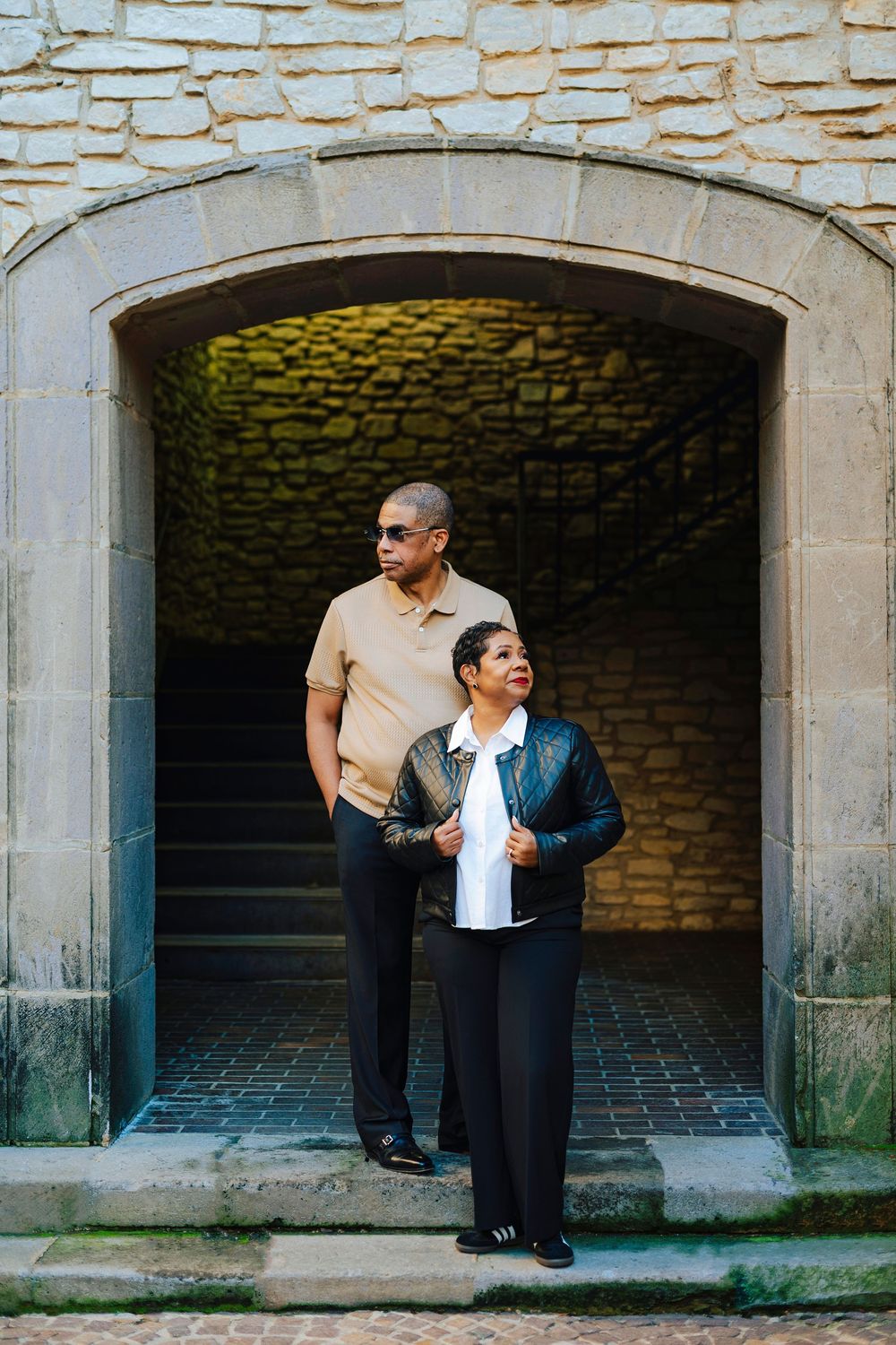 A couple stands in a stone archway, man in beige polo and sunglasses, woman in leather jacket, both in black pants.
