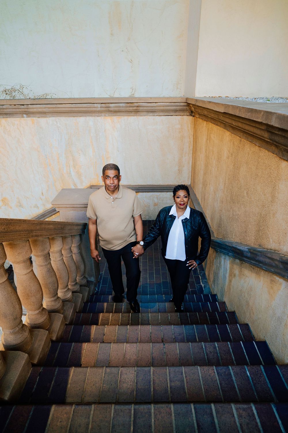A couple holds hands while descending an elegant staircase with stone balustrades and carpeted steps.