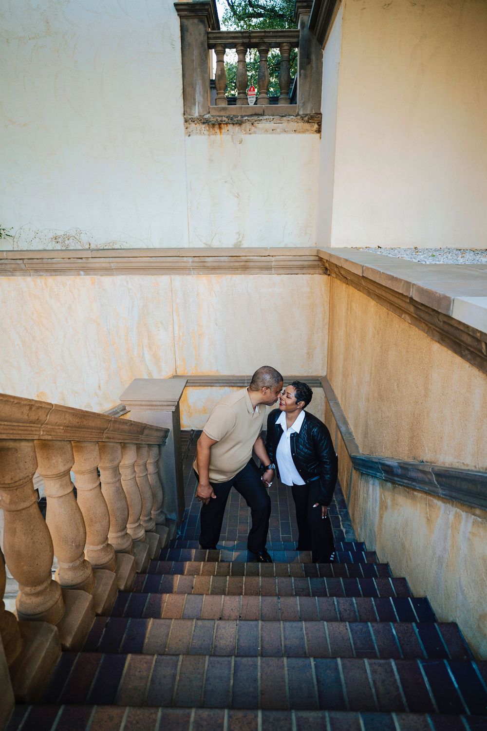 Couple holding hands on ornate stone staircase with balustrade, sharing a romantic moment at historic building.