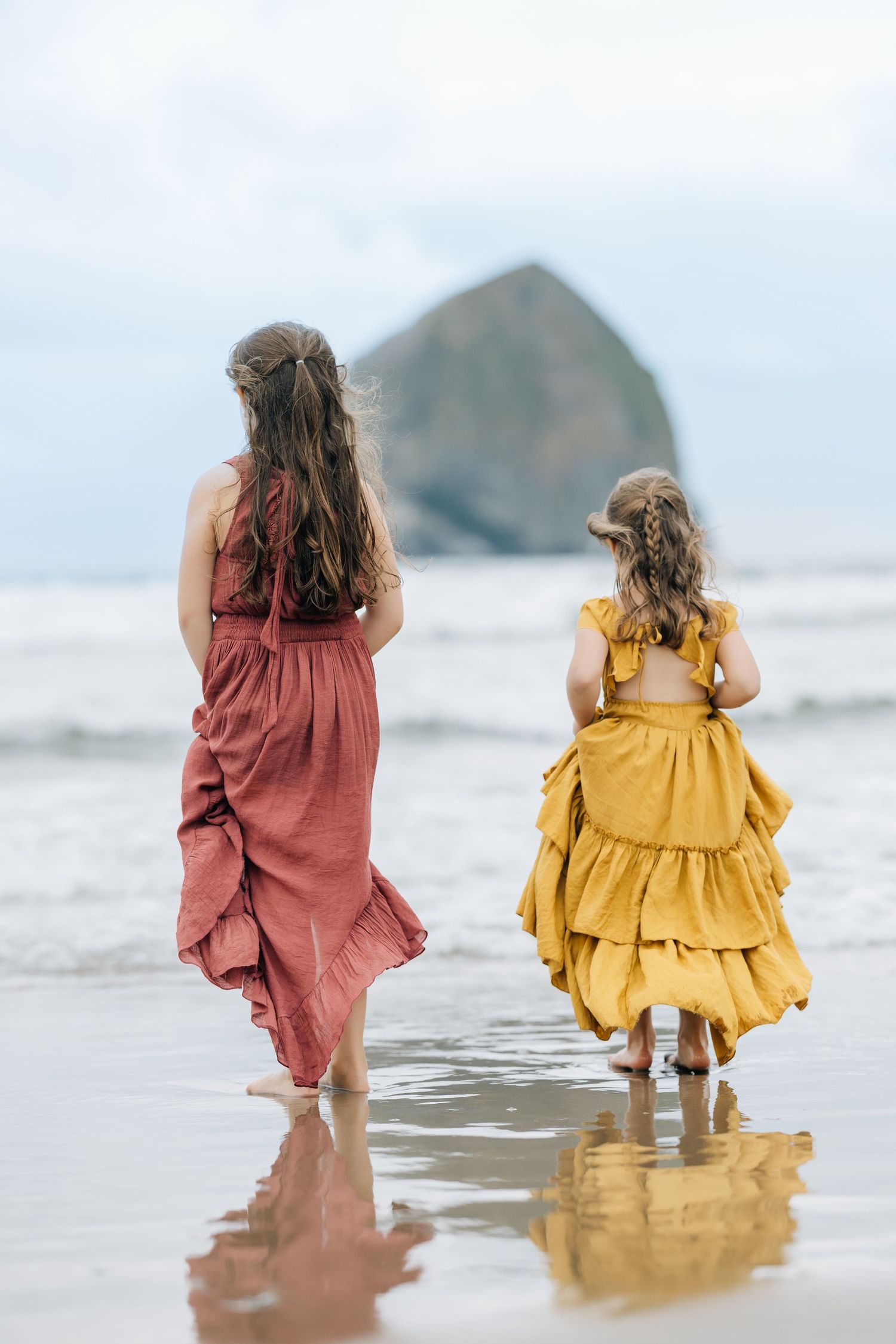 Two girls in boho dresses walk on a beach toward a large rock formation, their reflections visible on wet sand.