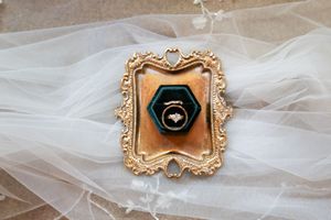 Gold ornate frame with emerald green hexagonal velvet ring box holding a diamond ring on a white veil.