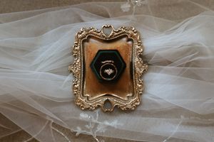 An ornate gold vintage frame holds a dark green velvet ring box with wedding rings displayed on a white veil.