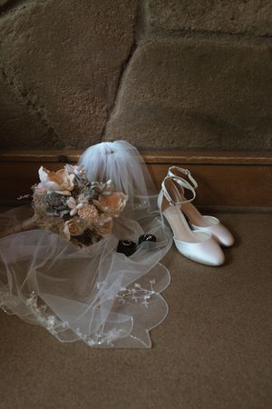 Elegant bridal accessories including white heels, floral bouquet, veil, and rings arranged on wooden steps.