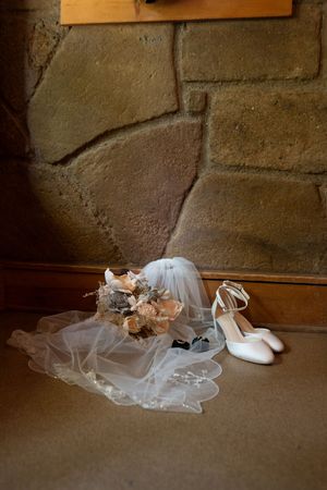Bridal accessories including white heels, floral bouquet, and veil arranged against a rustic stone wall.