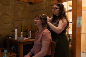 Hair stylist styling brunette womans hair in a rustic stone-walled room before a wedding ceremony.