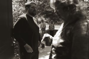 Black and white candid wedding photo of bearded groom with guests walking in background.