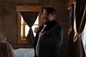 Man in black suit adjusts his tie while getting ready in a rustic stone-walled room for a wedding.