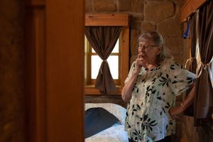 Elderly woman in floral blouse standing thoughtfully in rustic log cabin bedroom.