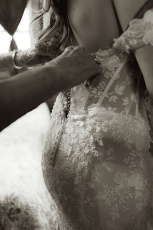 Black and white photo of a bride in lace wedding dress being buttoned up from behind.