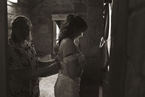 Black and white photo of mother helping bride into lace wedding dress in stone room.