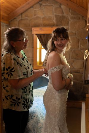 Older woman helps smiling bride button up her lace off-shoulder wedding dress inside a rustic stone cabin.