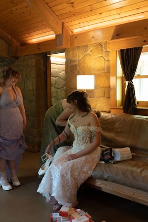 Bride in corset lace gown sits on leather sofa getting ready in rustic cabin.
