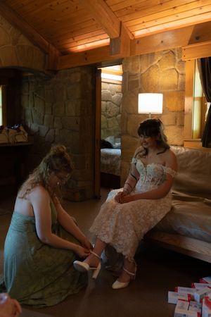 Bridesmaid helping bride with wedding shoes in warm wooden cabin with stone walls.