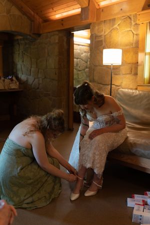 Bridesmaid in green dress helping bride with shoes in rustic stone interior room before wedding ceremony.