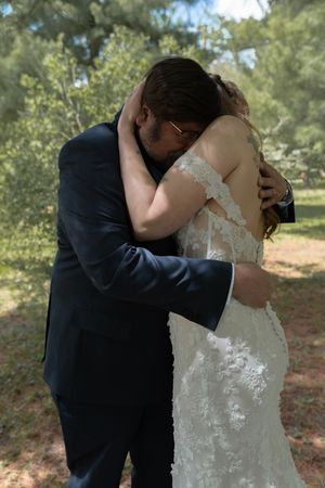 Emotional first look embrace between bride in lace gown and groom in navy suit outdoors.