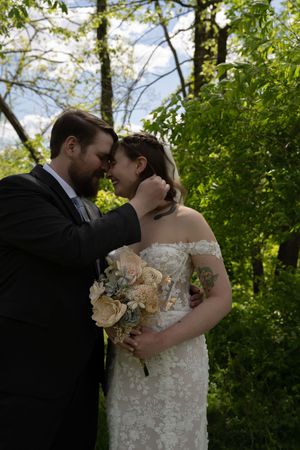 Groom tenderly cupping brides face as they smile together holding bouquet in sunlit green garden setting.