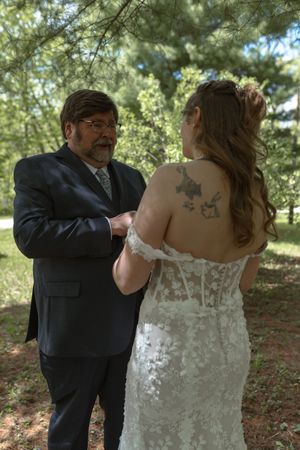 Father of bride fastening wedding dress buttons in sunlit forest setting.