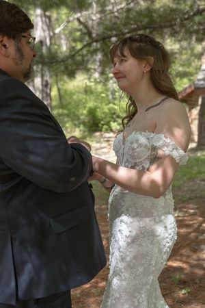 Bride in off-shoulder lace gown smiles joyfully while exchanging rings with groom in forest setting.