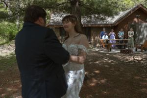 Bride in lace gown smiles joyfully during outdoor first dance near rustic cabin surrounded by pine trees.