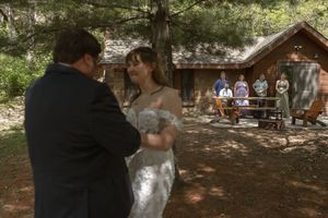 Bride and groom share first dance outdoors near rustic cabin with wedding guests watching nearby.
