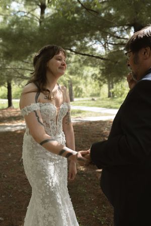 Radiant bride in off-shoulder lace gown holds grooms hands while smiling during outdoor woodland wedding ceremony.