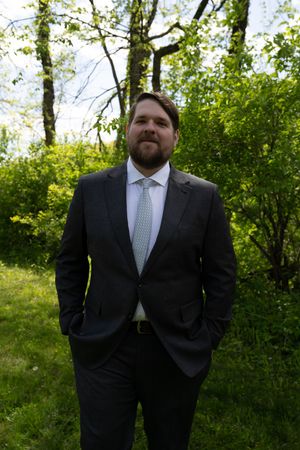 Bearded groomsman in dark suit and light tie poses outdoors among green trees and shrubs.