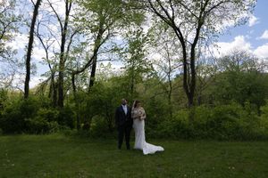 Bride and groom standing together in a sunlit green meadow surrounded by tall trees on wedding day.